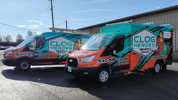 Two wrapped Clog Heroes service vehicles, a Transit van and a utility-body truck, parked side by side