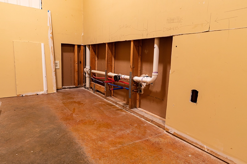 Room with the drywall cut away, showing white PVC drain pipes and red and blue supply lines running through the studs