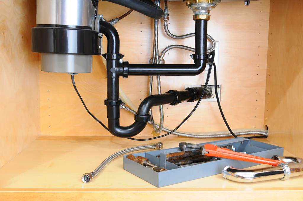 Black P-trap under a kitchen sink with a garbage disposal, and a pipe wrench in a tool tray