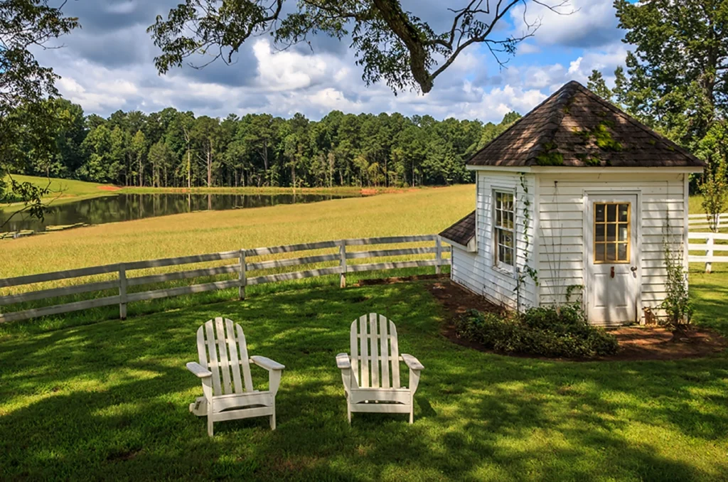 Small white well pump house with a shingled roof at the edge of a lawn with two Adirondack chairs