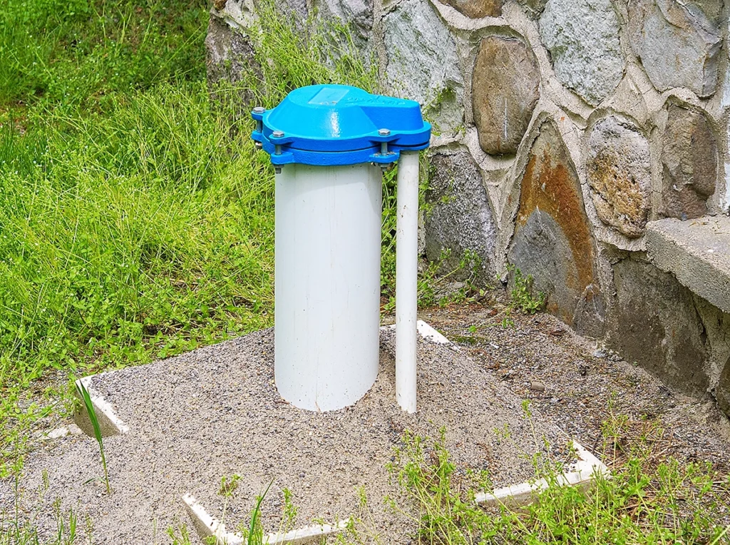 White well casing with a blue cap rising from a gravel pad next to a stone wall