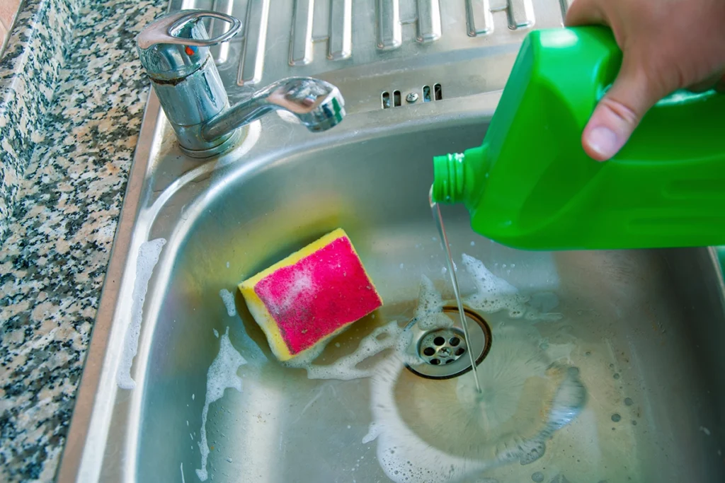 Hand pouring cleaner from a green bottle into a stainless steel kitchen sink drain